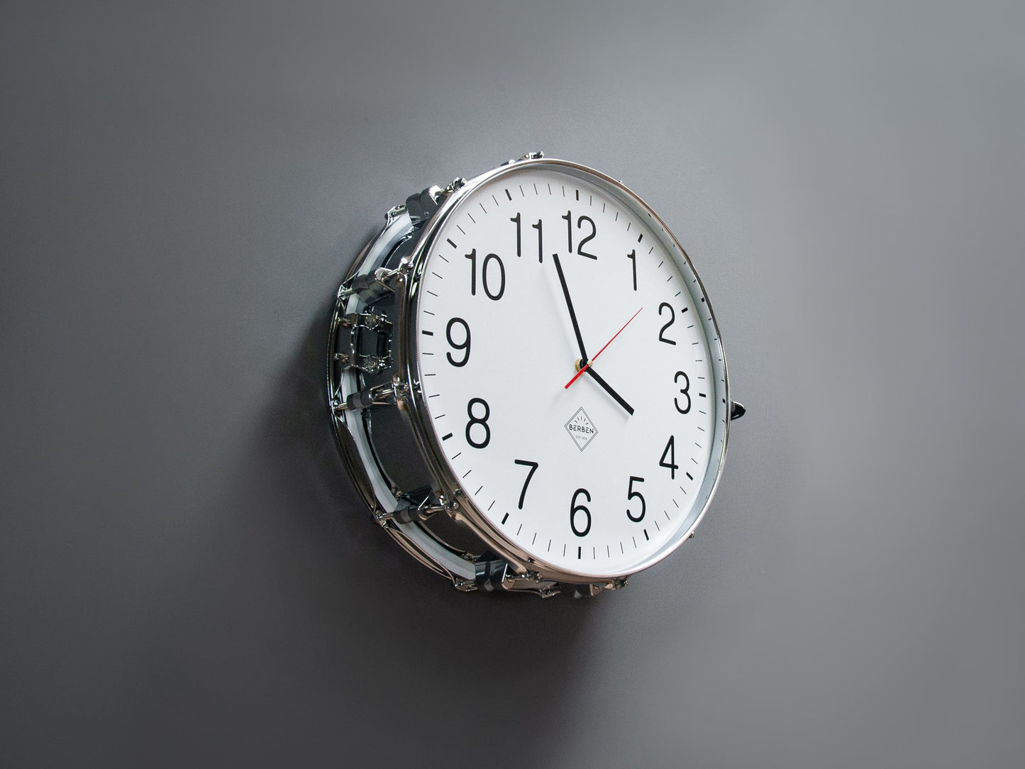 A clock made from a drum with a white clock face, mounted to a wall. The shell and rims have a chrome finish.