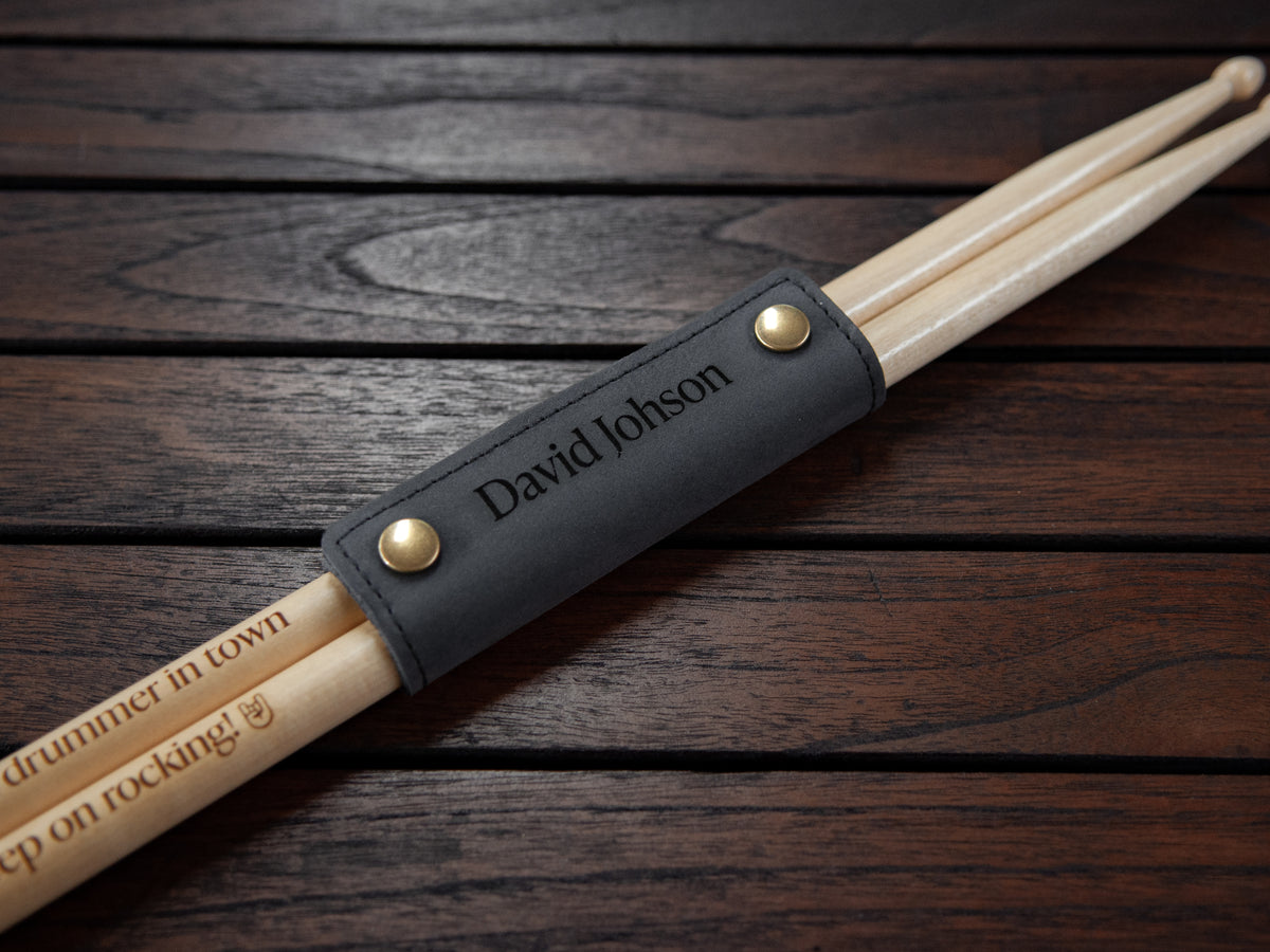 Personalized drumsticks in a black leather sleeve with gold snap buttons, engraved with the name 'David Johnson' and 'drummer in town', resting on a dark wood background.