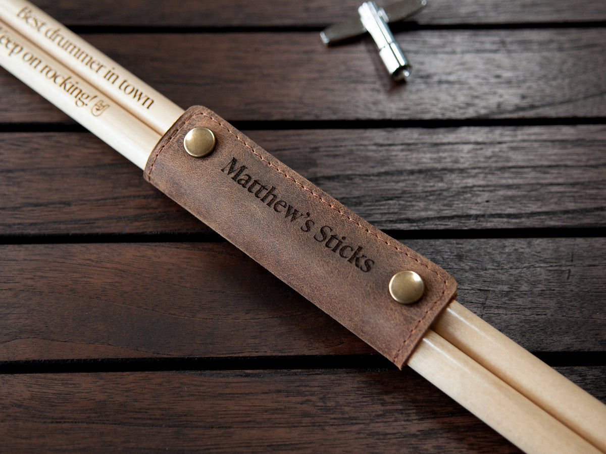 Personalized drumsticks in a brown leather sleeve engraved with 'Matthew's Sticks', displayed on a dark wooden background.